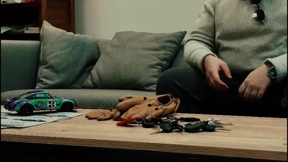 An enthusiast sitting on a sofa with driving gloves and a Porsche 911 model